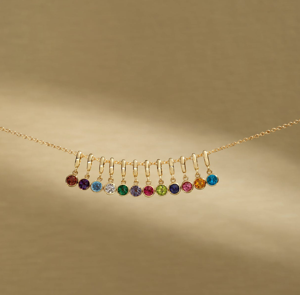 A delicate gold chain necklace featuring a curved bar of small, colorful gemstone charms in red, purple, blue, green, yellow, and orange, displayed against a warm, textured gold background.