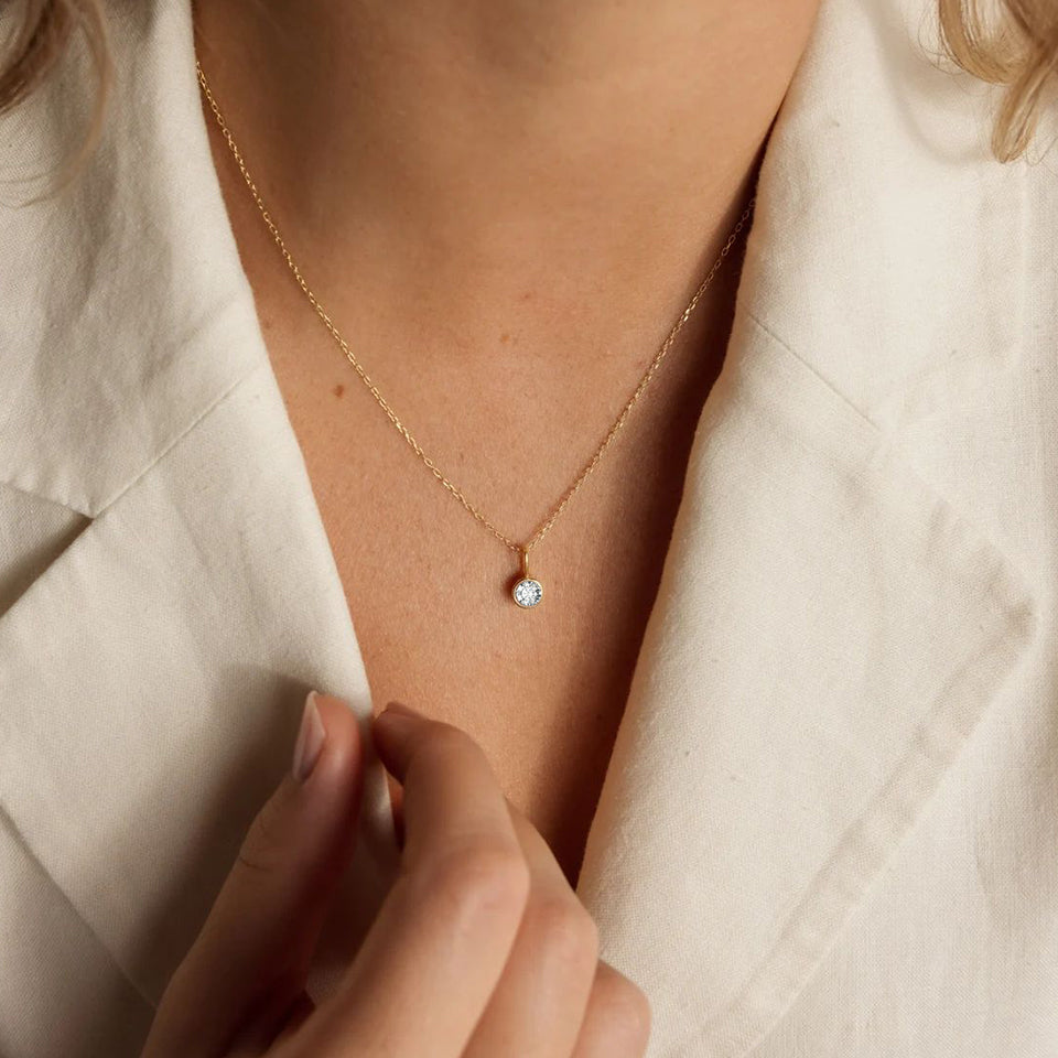 Close-up of a gold necklace with a small round pendant worn by a person in a light-colored shirt.