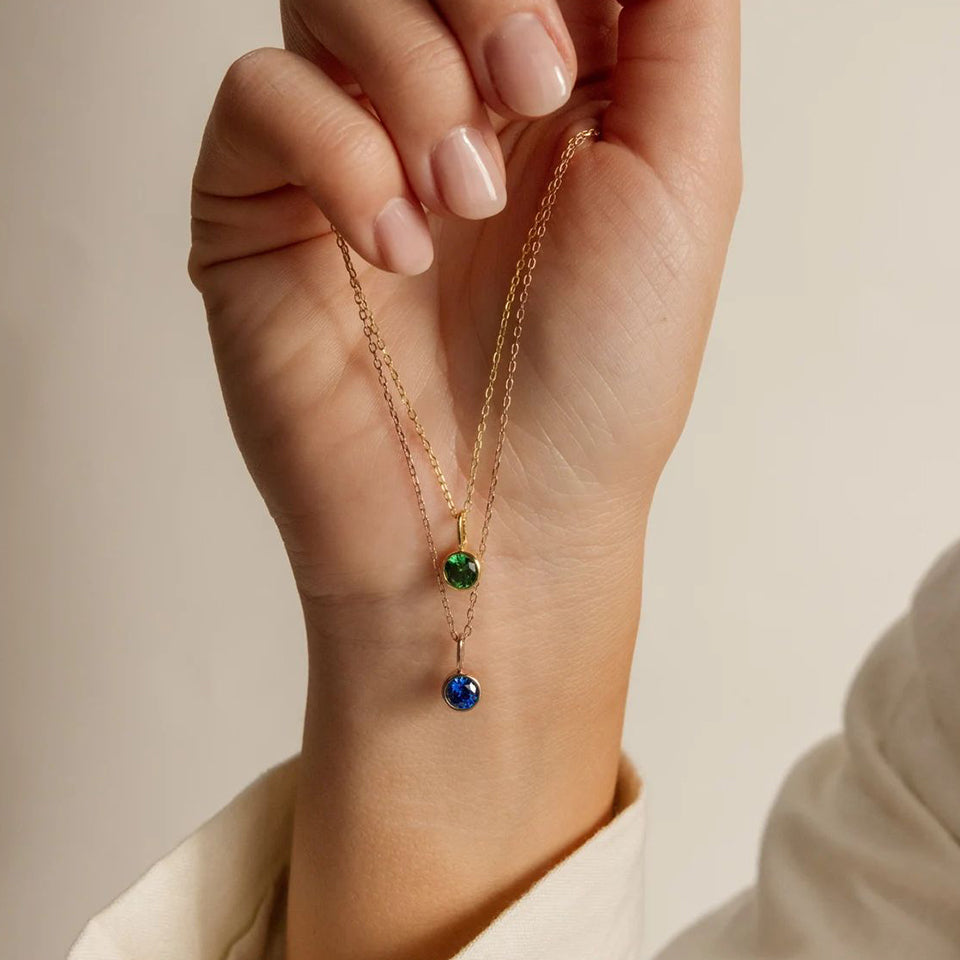 Hand holding a gold necklace with green and blue pendant against a neutral background